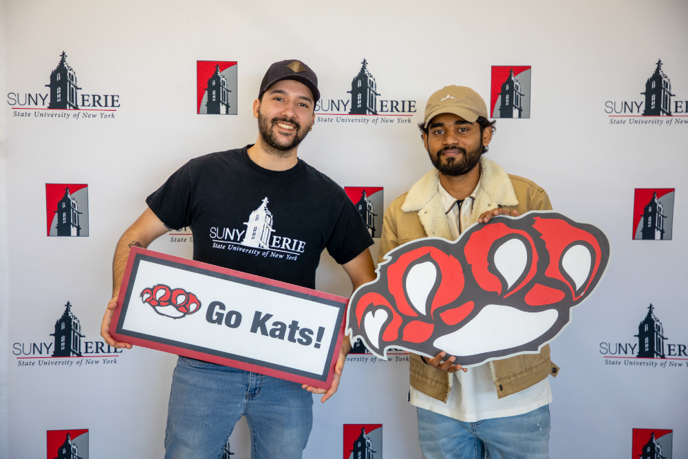 Two men stand in front of a step-and-repeat backdrop for SUNY Erie, smiling and holding a “Go Kats!” sign and a large red paw print cutout representing the school’s mascot.