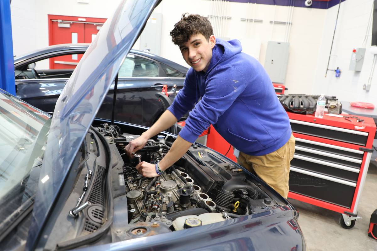 Student wearing a blue hoodie smiles while working under the open hood of a car in an automotive lab, using tools to service the engine, with tool cabinets and another vehicle visible in the background.