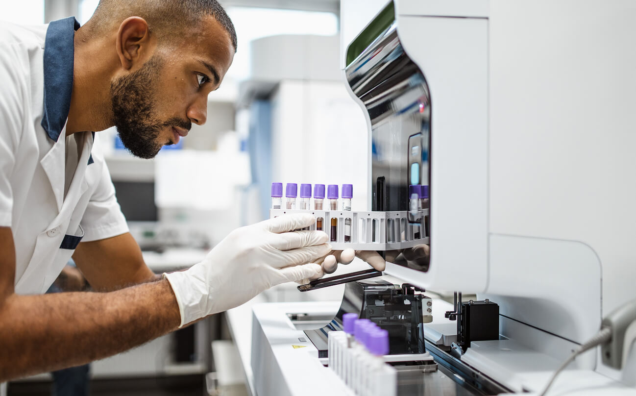 A clinical laboratory technician wearing gloves loads blood sample tubes into an automated laboratory analyzer in a modern medical testing facility. The technician carefully monitors the equipment as it processes patient specimens for diagnostic testing.