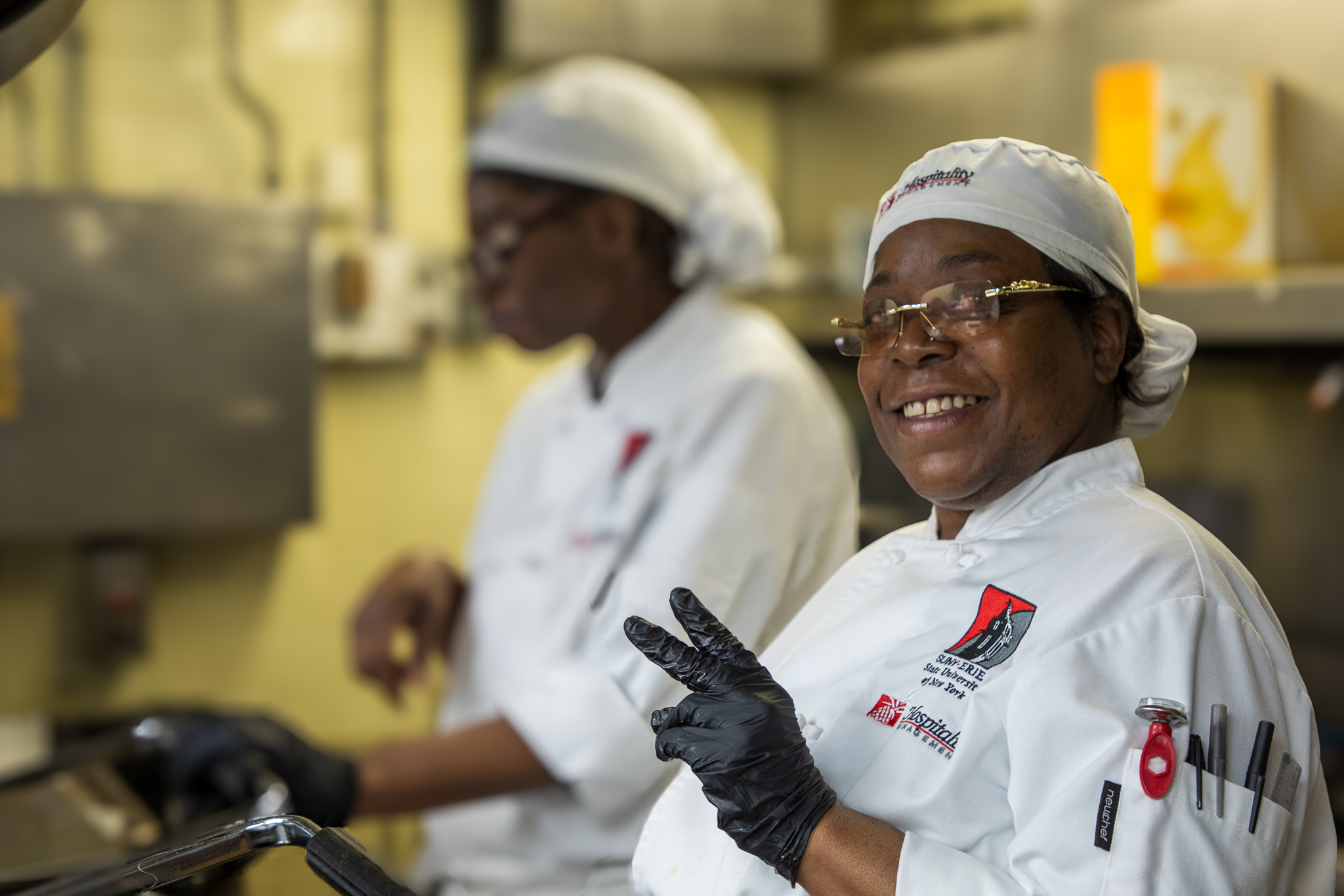 A culinary arts student wearing a chef’s coat and gloves smiles and flashes a peace sign while working in a commercial kitchen, as another student prepares food in the background at SUNY Erie.