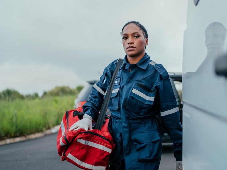 Paramedic walking next to her ambulance with a large bright orange bag over her shoulder.