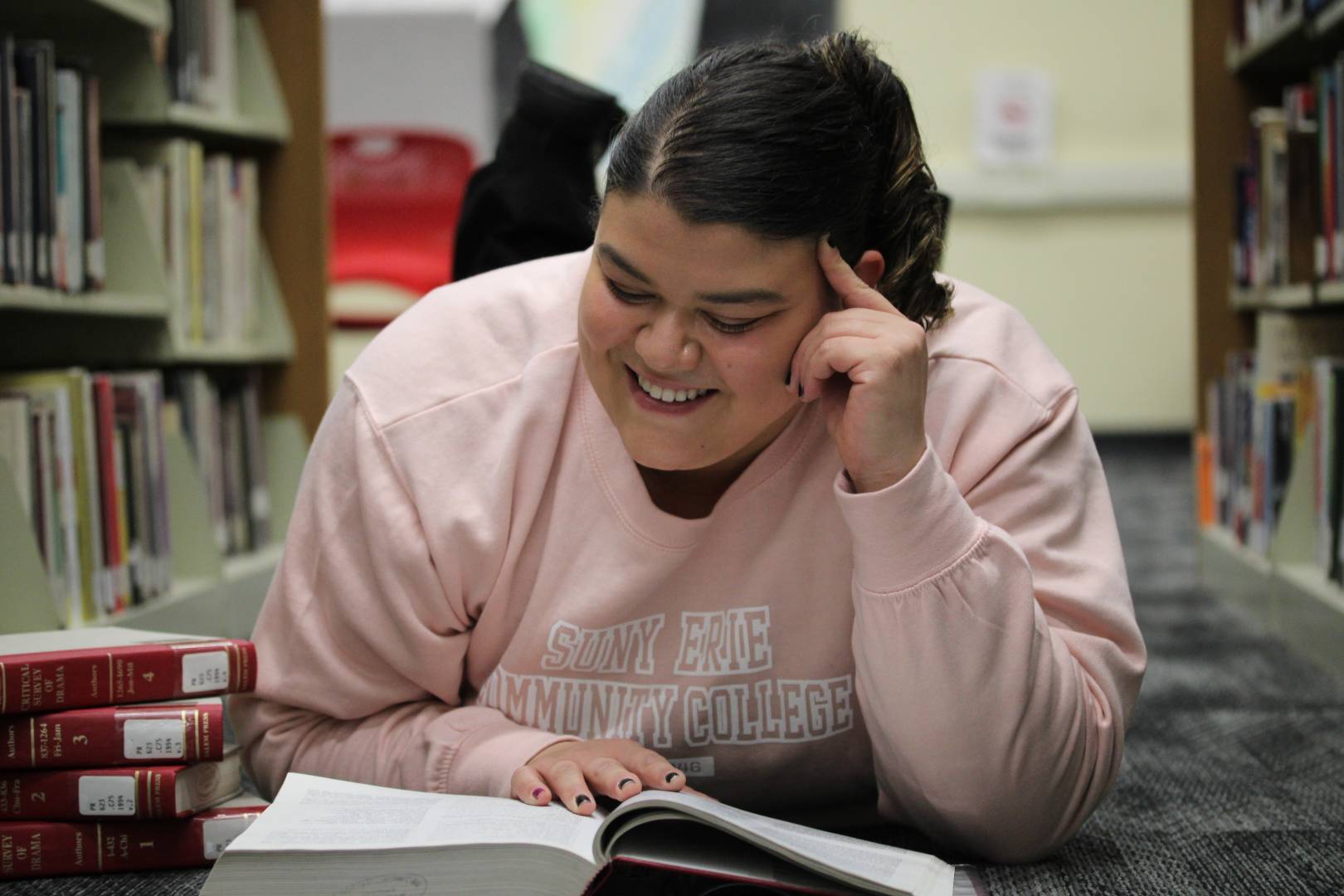 A student lies on the floor between library bookshelves, smiling while reading a book, with a stack of library books nearby,