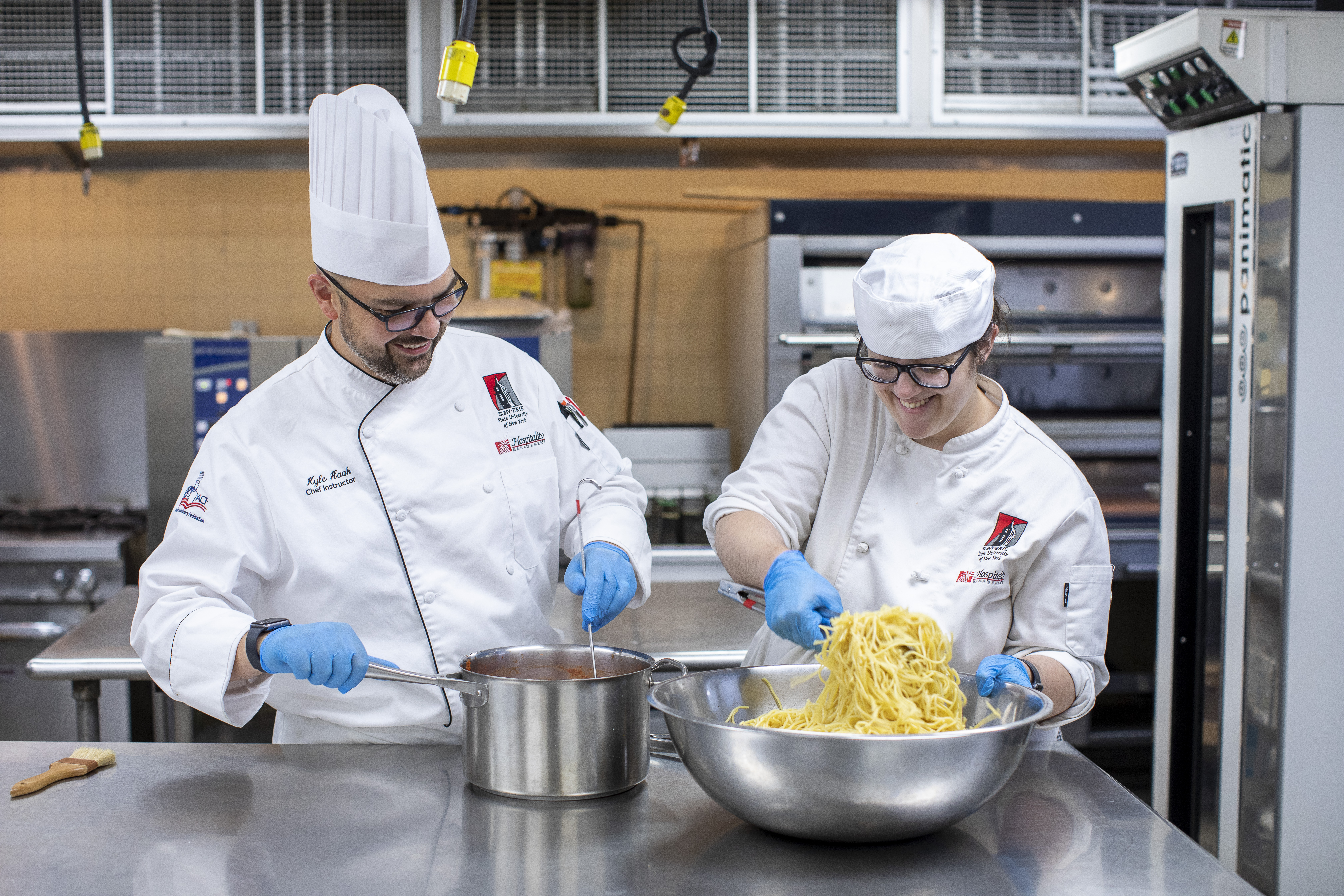 A culinary student and professor in chef coats and hats working in a commercial kitchen, the professor stirring sauce in a pot while the student other mixes freshly cooked pasta in a large metal bowl.
