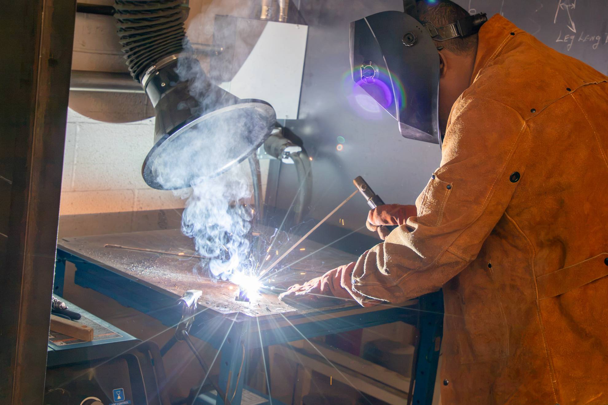 A student wearing protective welding gear and a leather welding jacket works with a welding torch on a metal fabrication project in a mechanical engineering technology lab.