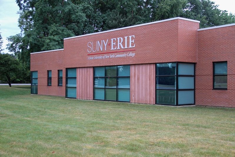 SUNY Erie Community College building with a brick exterior and large windows, viewed from across a grassy lawn with trees in the background.