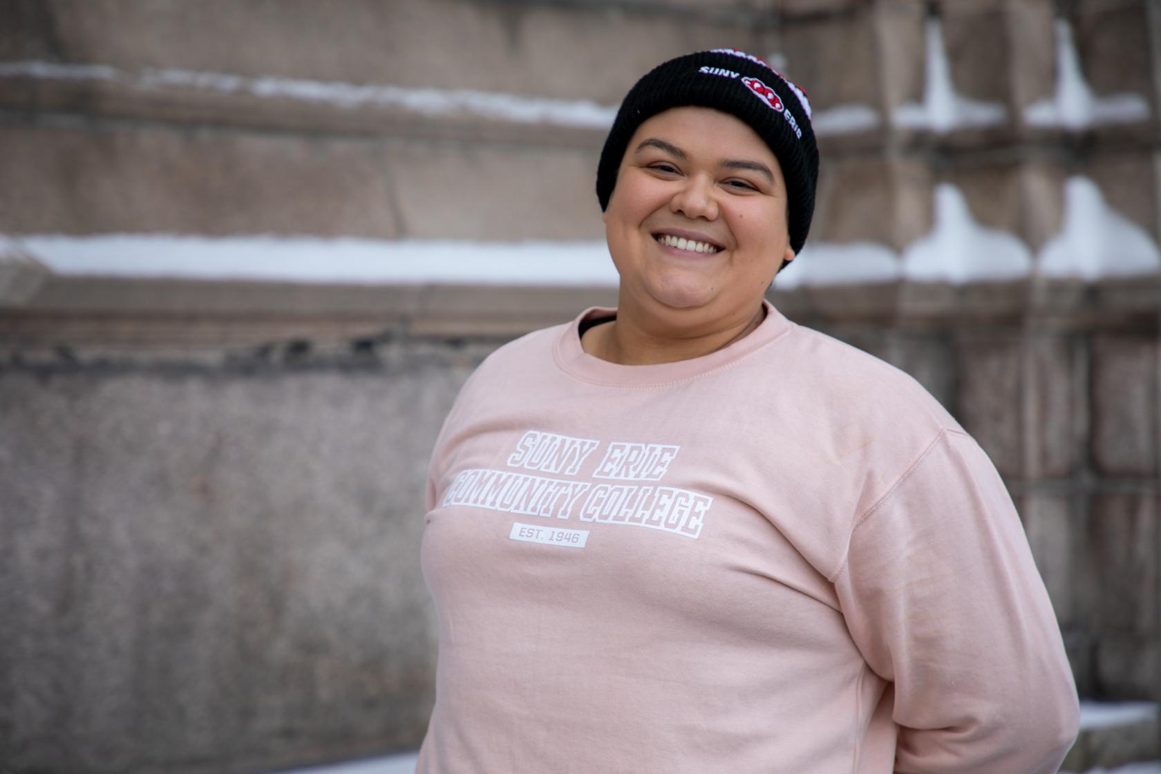 Smiling person wearing a black beanie and a light pink “SUNY Erie Community College” sweatshirt, posing against a snowy concrete wall.
