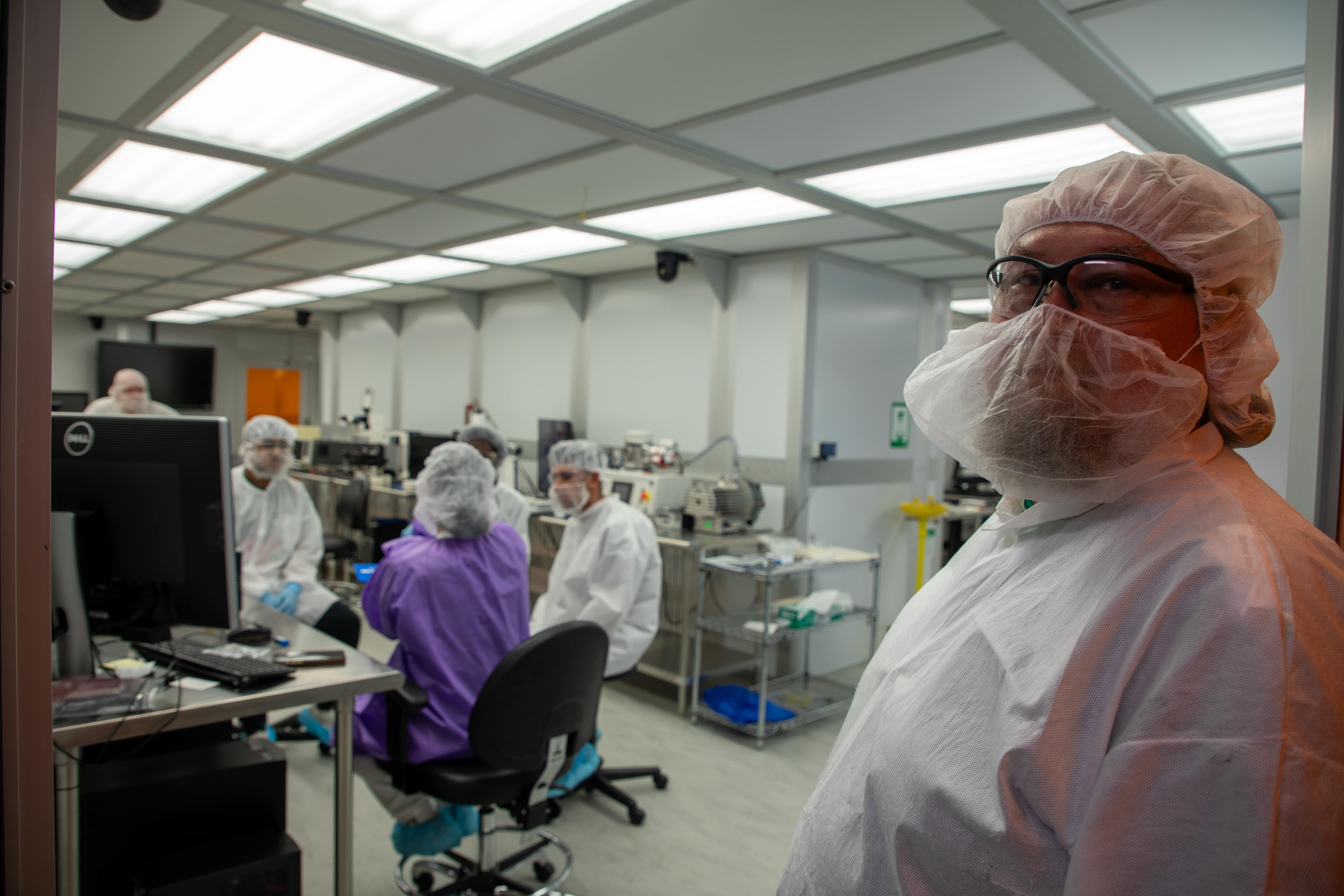 A group of people wearing full cleanroom attire, including gowns, hair covers, masks, and safety glasses, receive instruction inside a brightly lit laboratory filled with technical equipment. In the foreground, a person in cleanroom gear stands near the camera, looking toward the viewer while others sit and talk around a computer station in the background.